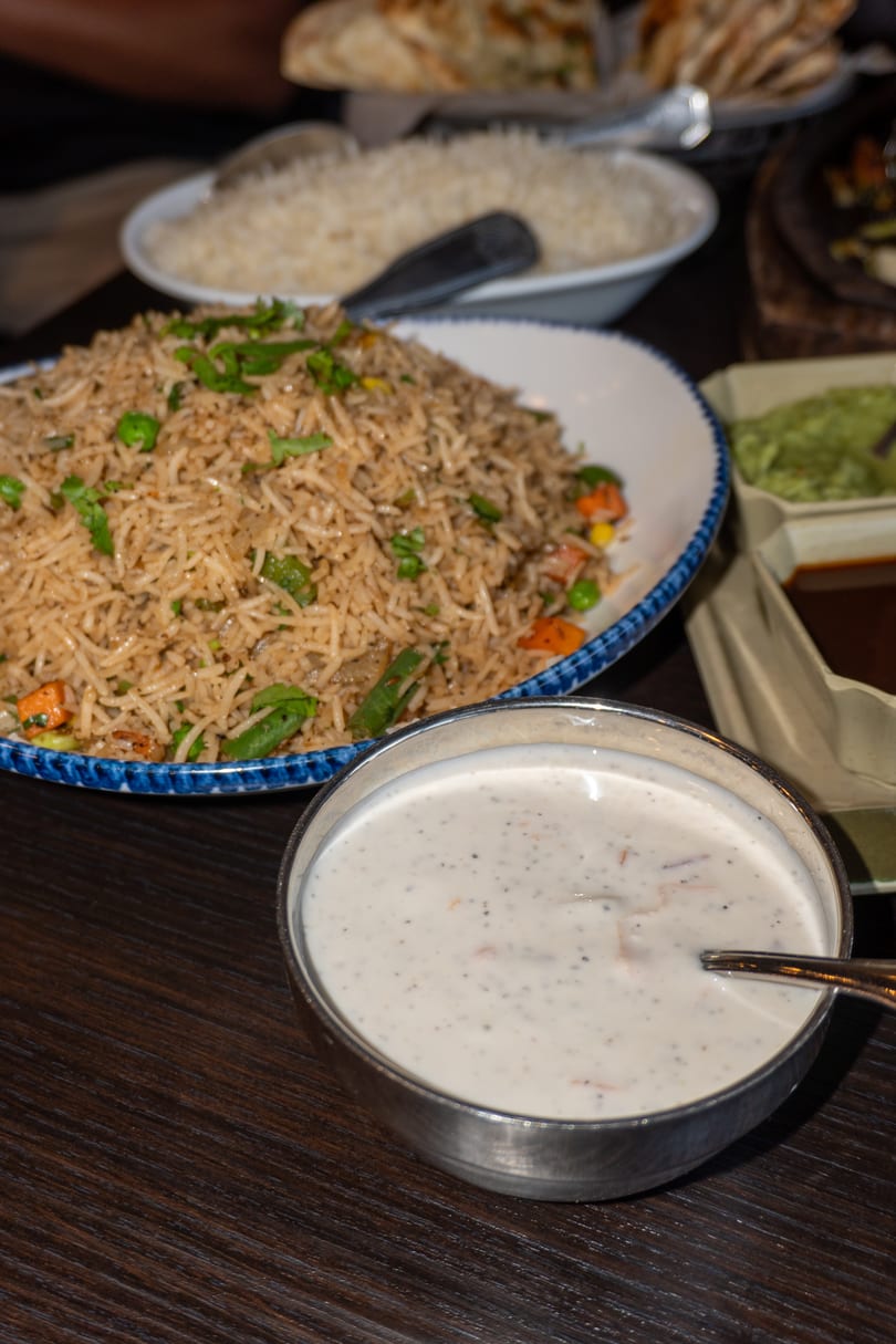 A bowl of raita with a spoon, next to a plate of vegetable fried rice on a wooden table.