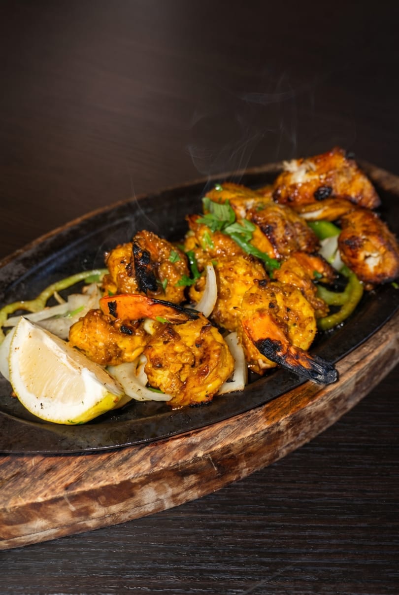Sizzling shrimp and chicken tikka with onions, peppers, and lemon on a dark wooden board.