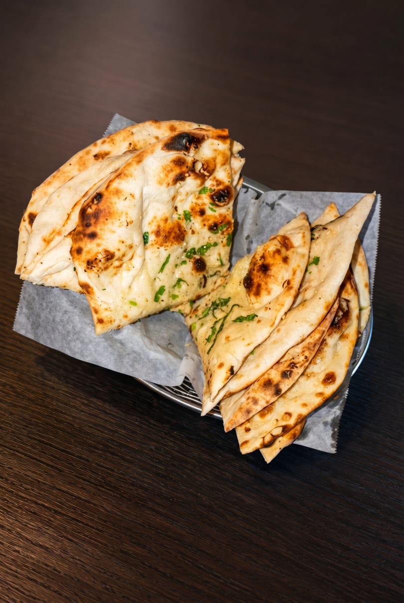 Two baskets of freshly baked naan bread, some topped with green herbs, on a dark wooden table.