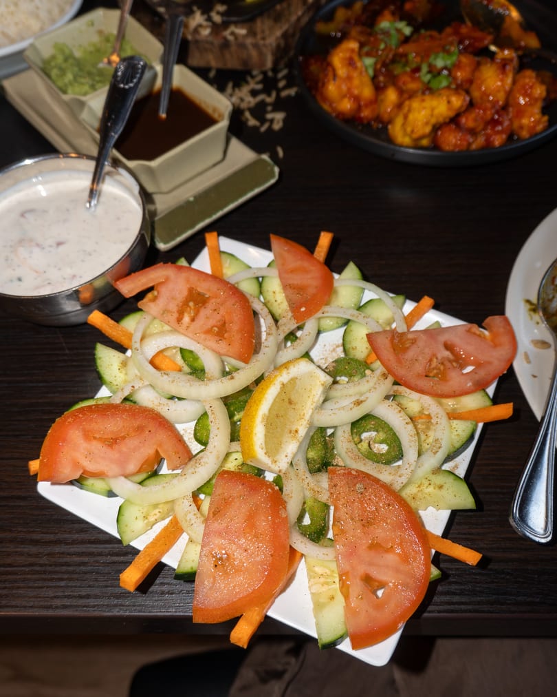 Fresh salad with sliced tomatoes, cucumbers, onions, jalapeños, and a lemon wedge, next to sauces and chicken.