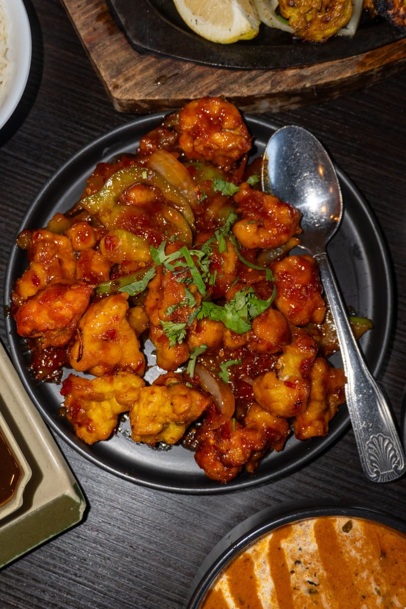 A black plate of orange-glazed chili chicken with green peppers and cilantro, with a spoon.