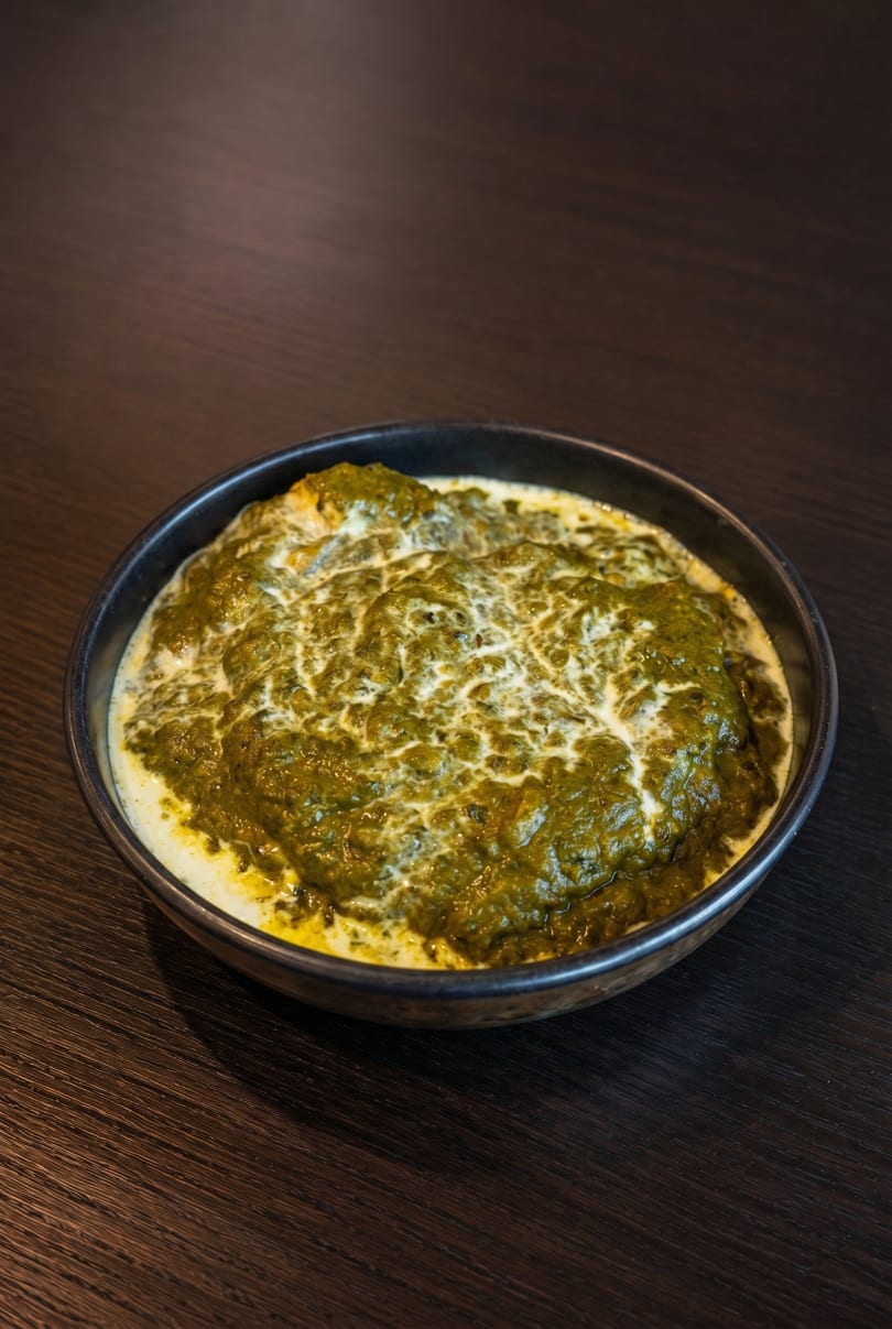 A dark bowl of vibrant green palak paneer with swirls of white cream on a dark wooden table.
