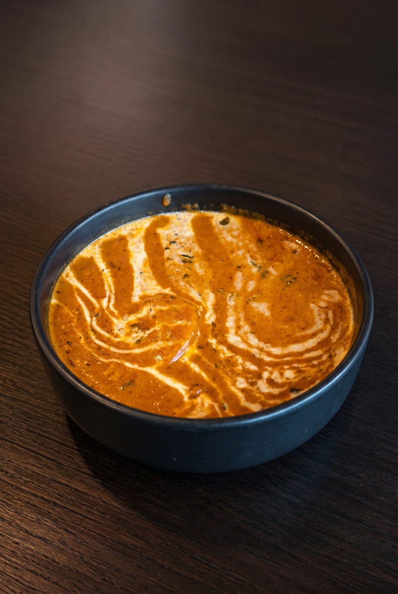 A black bowl of creamy orange curry with white swirls and herbs on a dark wooden table.