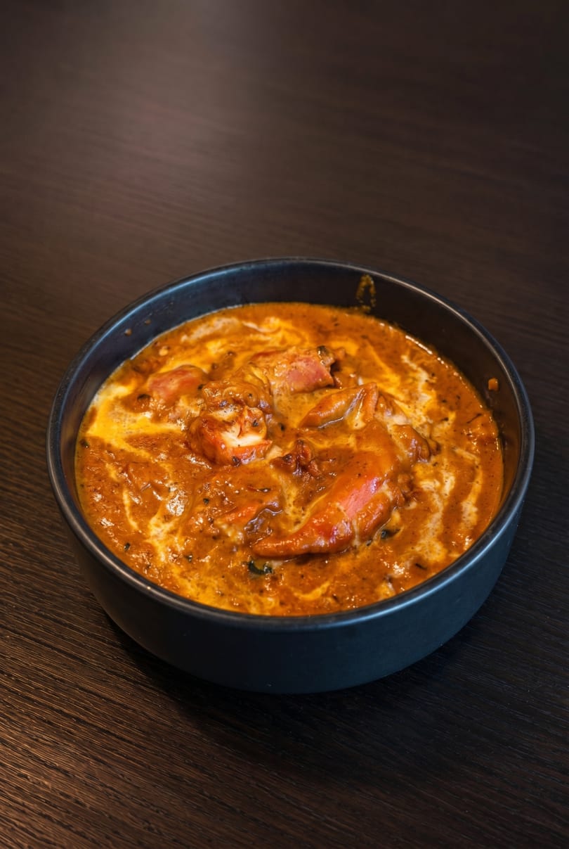A black bowl filled with orange curry, chicken, and a swirl of cream on a dark wooden table.